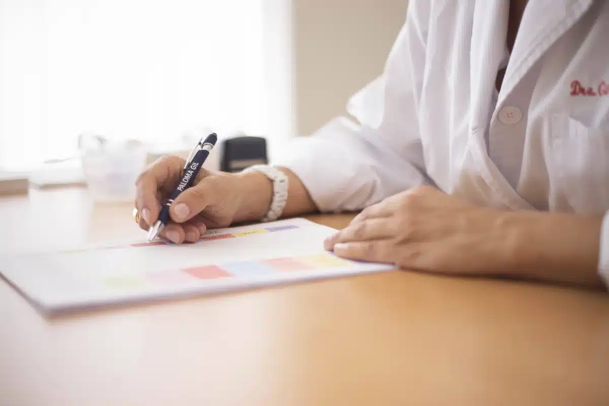 Mani di un caregiver con medicinali e documentazione sanitaria accanto, finestra chiusa con sole estivo