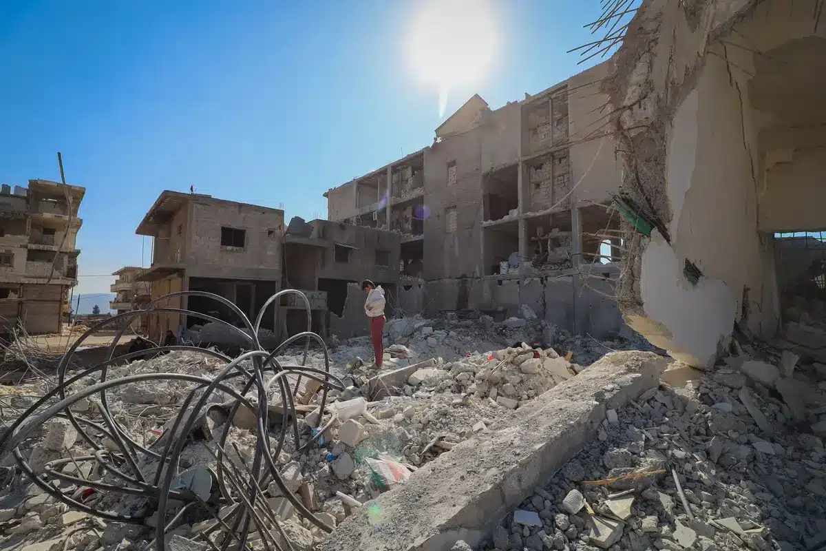 Strada distrutta con macerie, operatori di emergenza e civili tra le rovine di edifici danneggiati