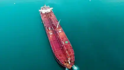 Petroliera in transito in uno stretto con navi militari sullo sfondo e terminali petroliferi costieri visibili