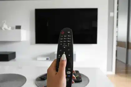 Una mano con telecomando puntato verso uno schermo televisivo spento in una stanza buia.