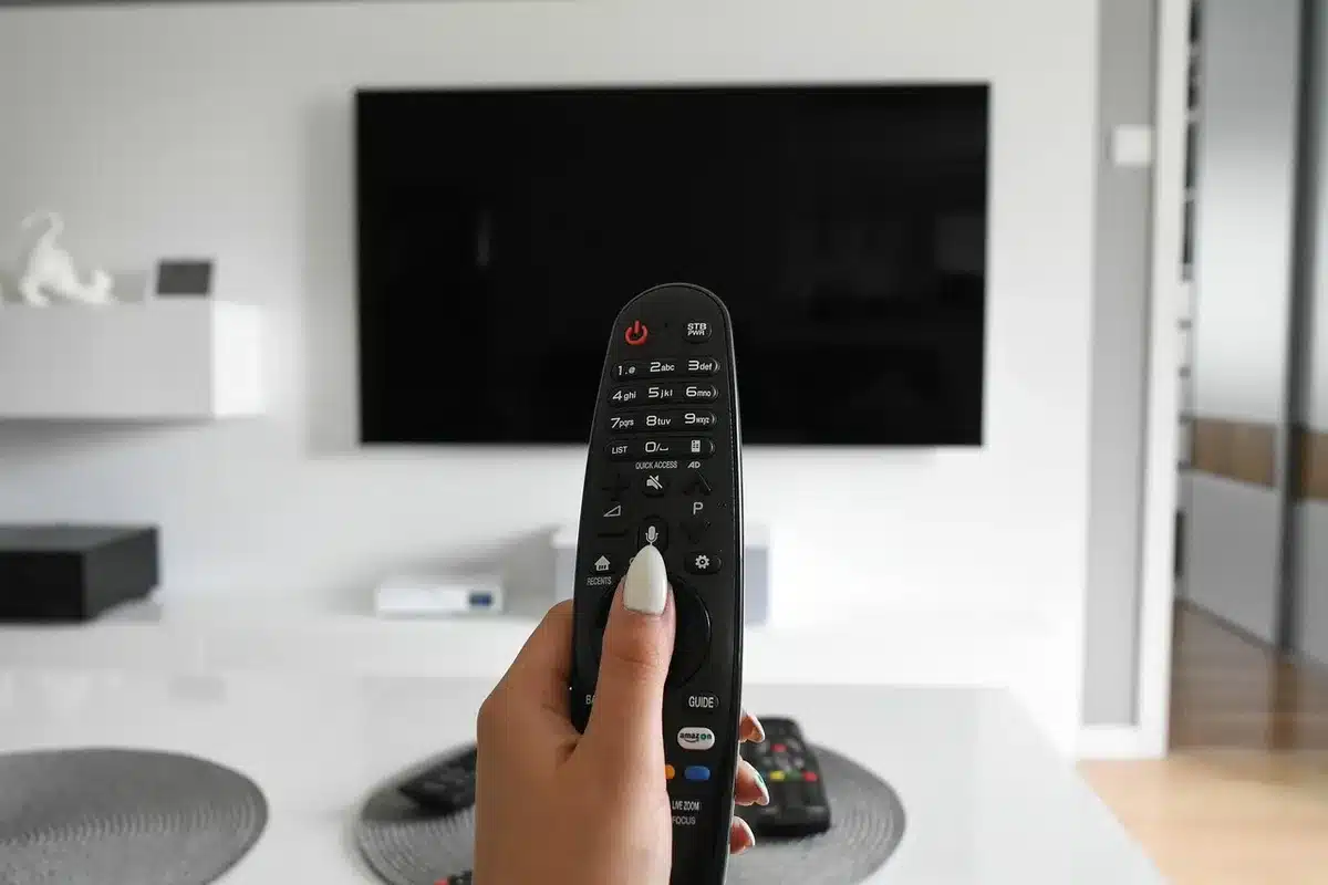 Una mano con telecomando puntato verso uno schermo televisivo spento in una stanza buia.